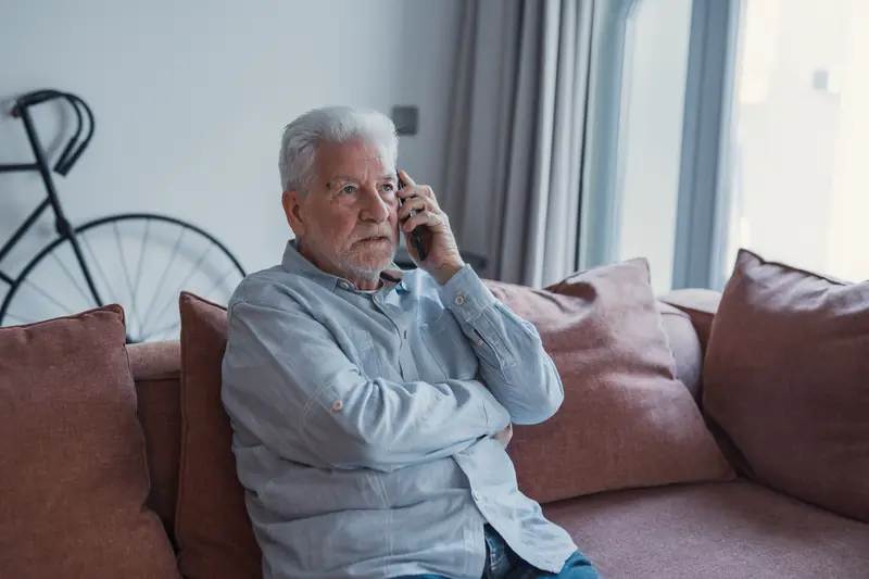 Älterer Mann telefoniert mit ernstem Blick auf dem Sofa in seinem Wohnzimmer
