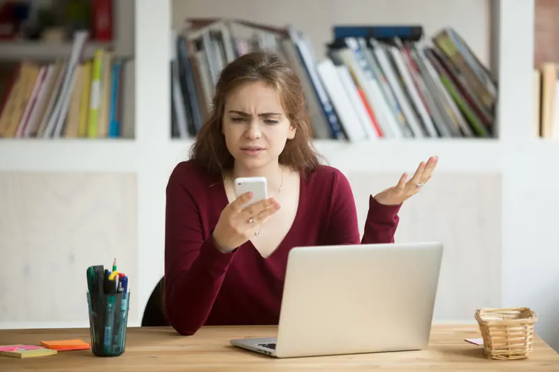 Frau sitzt am Schreibtisch, schaut verärgert auf ihr Handy und hebt fragend die Hand